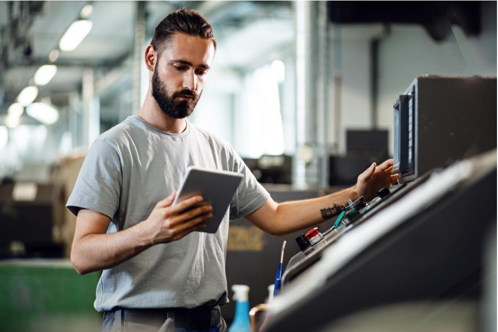 Man with a beard standing in a factory, holding a tablet while adjusting controls on a large industrial machine.